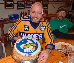 Craig sitting at the table holding his birthday cake with streamers on his head and to his left is Marty