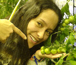 Patty holding tomatoes close to her face and pointing at the tomatoes while she holds a pollinating brush