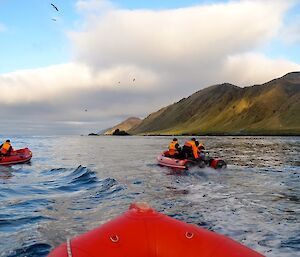 View from one of the boats (the bow in the foreground) looking south along the rugged coast, with sun illuminating part of the hills in bright shades of green. The other two boats are just ahead