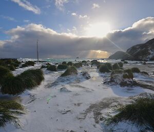 View towards North Head and the sun, low in the sky beaming rays through the top and bottom of the cloud. In the foreground iare windswept tussock mounds with icy snow covered ground in-between