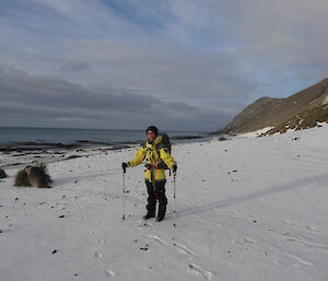 Patty dressed for the snow wearing a yellow jacket with her pack on holding both her poles standing in snow by the ocean with the mountain hillside.