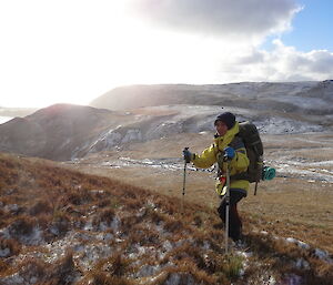 Patty dressed for the snow wearing a yellow jacket with her pack on holding both her poles standing with the snow covered mountain range behind her