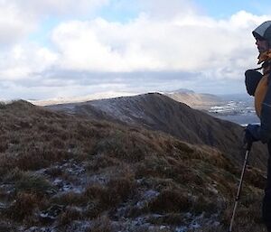 Marty facing east dressed for the snow with his hood over his head wearing his pack and holding his pole with the mountain range and ocean fluffy clouds and blues sky.