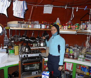 Patty holding a kettle at the kitchen wearing a scarf that goes to her chin making tea