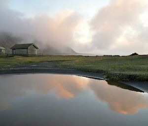 A photo of a puddle reflecting the colures of the sky which are soft pink, blues and greys and in the back ground are huts amongst a tussock hill