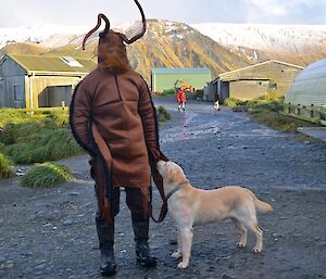 Steve Horn (dressed in a cockroach costume) bringing his charge "Flaximus Prime" (white labrador) to the race course. snow covered hills can be seen in the background
