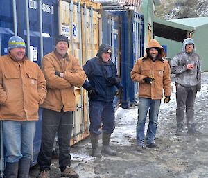 The enthusiastic crowd, of five epeditioners, awaiting the start, while sheltering behind some shipping containers out of the wind, cold and snow