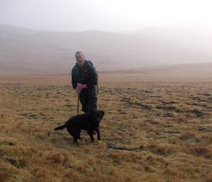 Nick with Wags, a black labrador, standing on some grassy field with the hills rising in the misty background
