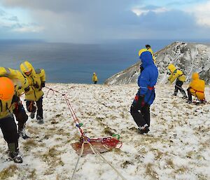 Rescue team working to raise the stretcher — on a slight snow covered slope above Gadgets Gully. Three members of the team haul on the rope and pulley system, while the team leader relays hand signals to a spotter at the edge of the steep slope and to the right is a two person belay team