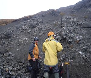 Marty and Josh stand just below the leaking pipe on the scree slope, assessing and making preparations to fix the leak. Both are wearing safety helmets