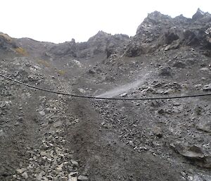 Looking up the scree slope of Gadgets Gully with the black poly water pipe running across the slope, slightly suspended above the surface. Water can be seen spraying from a leak in the pipe