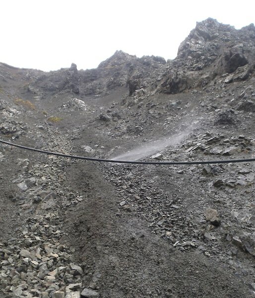 Looking up the scree slope of Gadgets Gully with the black poly water pipe running across the slope, slightly suspended above the surface. Water can be seen spraying from a leak in the pipe