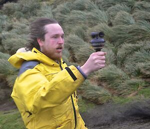 Aaron getting a reading of the wind alongside Razorback ridge. His hair is swept back and his cheeks puffed and he is holding a hand held anemometer