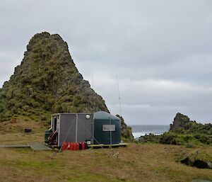 A water tank hut with an annex, aerial, decking and fuel containers on a grassy area with rock stacks and the west coast in the background