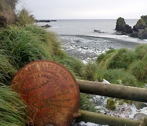 Old water cart lid leaning against a pine fence next tussocks with the green gorge coast line in the back ground