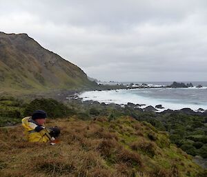 Ranger Chris relaxing on a grassy knoll while looking down the rugged west coast with the escarpment on the left half and the surf on the right half