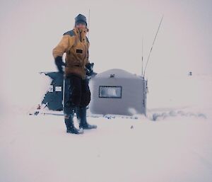 Ange standing on the snow near the exposed metal hook of the cage pallet she is about to dig out. Eitel hut, a converted water tank, is in the background