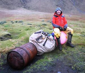 Aaron sitting on a large fishing float which is next to a big bag and a large metal drum which is all part of a cache of marine debris established at Sellick Bay in 2012