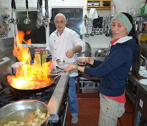 Patty and Chef Tony in the kitchen, preparing the food for Dave’s party. Patty is holding a large frying pan over the stove top that has flames rising from it