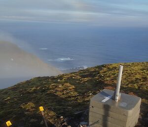View from the top of Mt Waite. In the foreground is a concrete block which is support for the antennae. The slopes below have a light mist and surf can be seen breaking on the coastal rocks far below