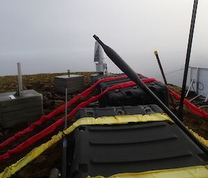 The Mount Waite snapped antenna shown leaning against a large plastic storage crate which is heavily strapped down. The concrete base for the antenna can be seen behind the crate. A wind turbine as well as a solar panel are also in the photo