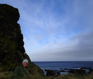 Josh, at the bottom of the picture, amongst the tussock, next to a large rock stack, with the calm ocean in the background