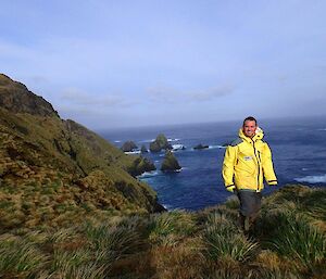 Josh standing amongst the tussock with the steep rugged slopes of the escarpment in the background. There are also some impressive large pointy rock stacks in the ocean just offshore