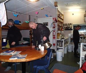 Inside Hurd Point hut — shows ranger Chris working in the kitchen, while Craig can be seen pouring water from a kettle into a mug on the oval table. Clive is standing behind the table