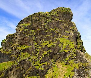 A large rock stack covered in patches of moss, outlined by the beautiful blue of the sky. There is patches of high, white cloud