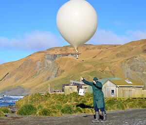 Craig, dressed in all the protective safety clothes, including goggles and gloves, releasing the large weather balloon with a ozone sonde attached. It is a bright sunny morning, highlighting the colours of the escarpment in the background