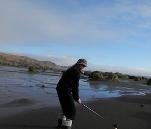 Angela Newport grasping a golf club (wood) addressing a yellow golf ball on the dark sands of Bauer Bay beach