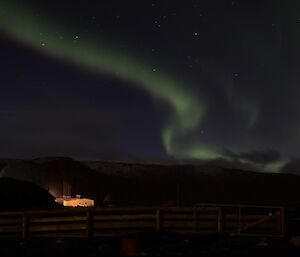 Aurora to the south of the station, in the shape of a elongated two, with stars seen in the background and the snow tipped escarpment silhouetted. The science building can be seen illuminated by a light from another building