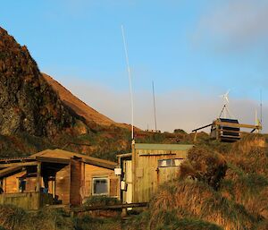 At sunrise, has an orange tint to it, the view of the rustic wooden hut and shed that is Green Gorge hut, on a slope nestled in long tussock like grass and large hills in the background