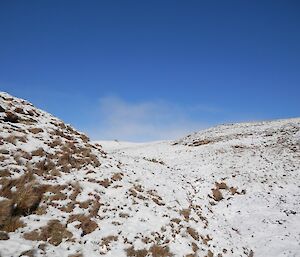 Blue skies after the big snow dump — looking up a slopping valley which is covered in deep snow, though some tussock grass mounds are sticking up out of the snow. A stark contrast with the clear blue sky