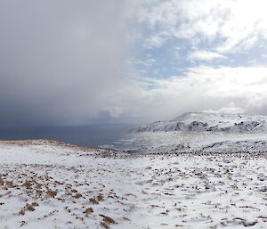 Another panorama taken from a high point on the plateau looking over a deep snow covered scene of the plateau in the foreground, a wide valley and the coast below, then the snow covered hills behind the valley. A snow shower can be seen moving across the ocean
