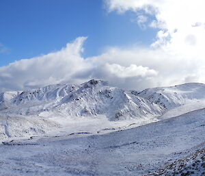 Panorama view taken from high up of a creek valley and the rugged slopes on the other side of the valley. The whole landscape is covered in deep snow and the and the dark blue ocean can be seen in the left of the scene