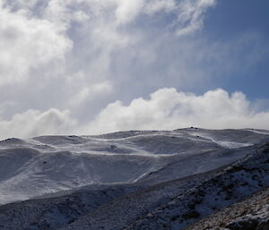 The rugged slopes covered in deep snow. Streaks of snow can be seen blowing near the peak of the ridge, with sun-drenched white cloud just poking up from behind the ridge