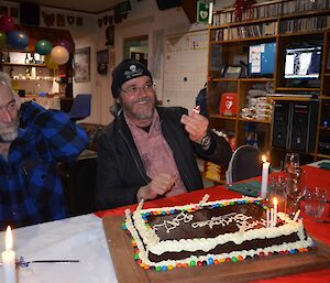 Barry sitting at a table with his big rectangular birthday chocolate mud cake. He has just let off a party popper and Lionel(sitting next to him) has his hands over his ears