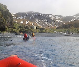 Arriving at Green Gorge — the red bow of a boat is in the immediate left foreground while another boat is just about reach the beach in front of Green Gorge hut. There are a couple of elephant seals and dozens of king penguins on the beach, while the escarpment in the background is covered in patchy snow