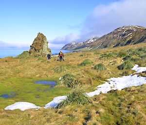 Mark and Greg carrying bags of dog food back from Waterfall Bay Hut — they are walking over an area of grass and tussock with some patches of snow and a single tall rock stack behind them. The east coast and rugged slopes of the escarpment are in the distant background