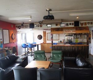 Bar and lounge — two couches and four single lounge chairs are around a large coffee table. In the background is the bar with several blue bar chairs and a dart board on the left wall. Over the bar is an old sign recovered from the old Caroline Cove hut — On the sign ‘Caroline Cove’ and ‘Crabbe Inn'