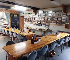 Dining area with three long dining tables and chairs. On the far wall are the photos of all past Expeditioners. An old clock is also on the wall with the kitchen bar visible on the left