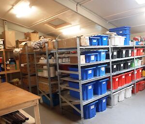 Field store and excess kitchen supplies in the MPB (Multi Purpose Building). There are several rows of shelving stacked with plastic bins and other items