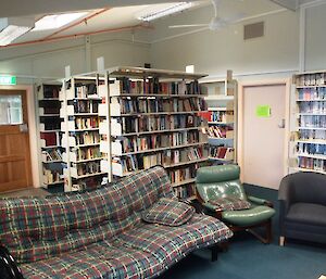 Library and sitting room in the MPB (Multi Purpose Building) — shows a lounge settee and two single lounge chairs with three shelve units stacked with library books. On the the wall behind are more shelves stacked with videos and dvd's