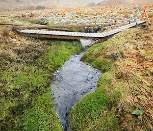 Broken bridge across Sawyer Creek near Green Gorge. The wooden slatted narrow bridge is bent at and angle over the creek where the timber supports have been broken