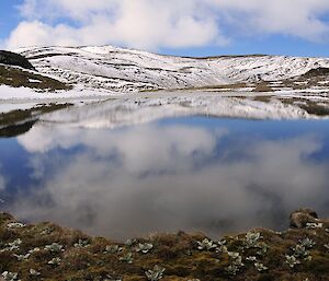 A beautiful image of Emerald Lake with an almost mirror water surface. In the foreground is the lake shore adorned with a variety of vegetation, while behind the lake are snow covered hills, which are reflected on the lakes surface