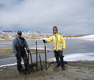 Chris and Clive removing old, rusted steel pickets at Island Lake. They are standing on the sandy shore of the lake with the snow covered hills in the background behind the lake