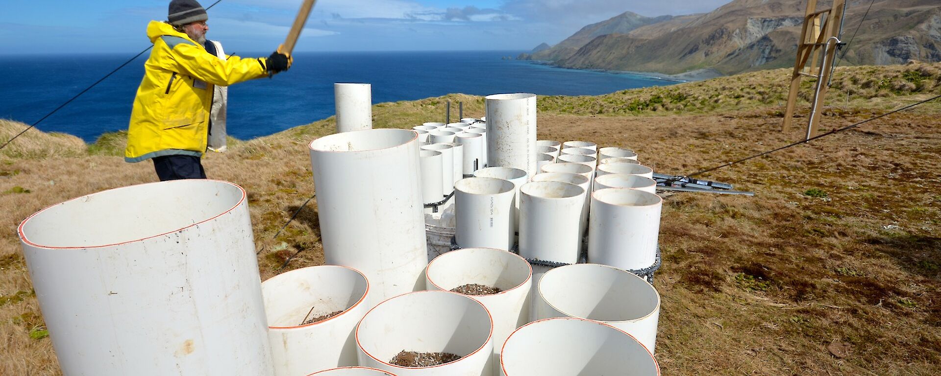 At the Wireless Hill Orchard site — Clive, using a piece of timber to hammer down and secure the final tubes into place within the steel frames. The east coast and ocean provide a scenic backdrop