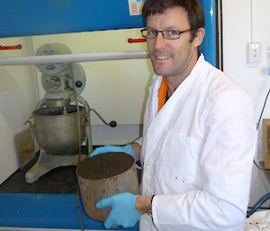 Grant, wearing a white laboratory coat is creating mesocosm samples (he is holding a sample in a tin). He is using the mixer on the bench behind him
