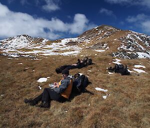 Jaimie and Marty taking a break at Cape Star. They are both laying against their packs on a grassy slope with snow covered hills in the background